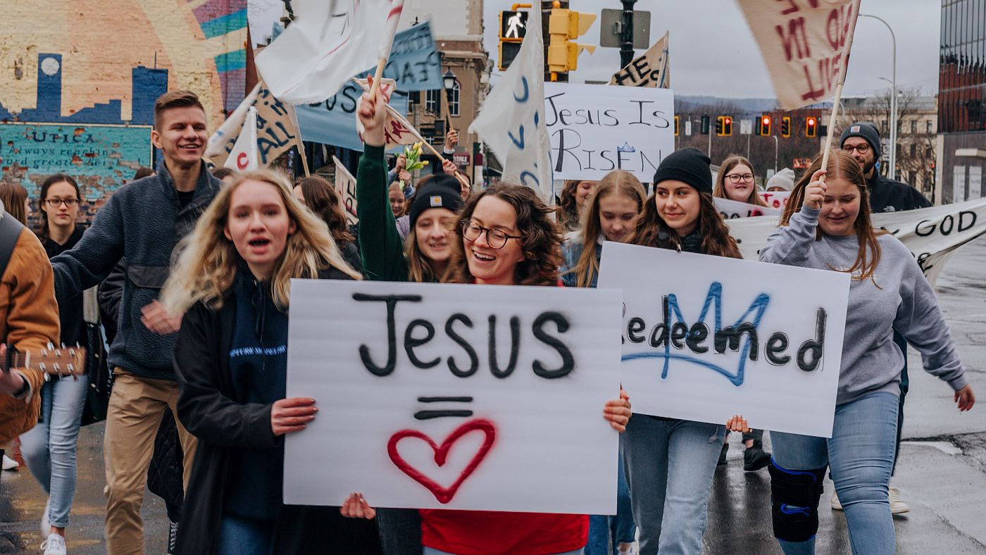 Jézus neét viselő transzparenssel menetelő vidám emberek
